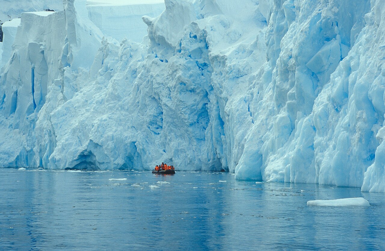 A zodiac boat carrying passengers in orange jackets between towering blue glacier walls